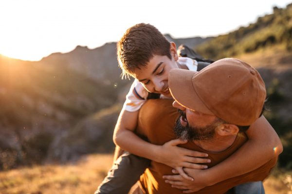 Cheerful dad carrying his teenage son on his back in nature on sunny summer day